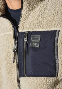 Fleece jacket featuring a cream body with navy accents, zippered chest pocket, textured fabric, and contrasting stitching details.