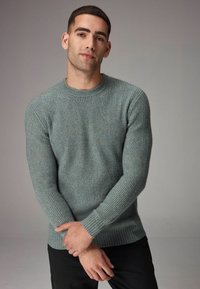 Pulover tricotat de culoare verde-albăstrie deschisă, cu textură ribbed, guler rotund și mâneci lungi, purtat peste pantaloni negri. Mâinile îmbrățișate în față.