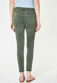 Kobieta nosząca dopasowane oliwkowe spodnie cargo z tylnymi kieszeniami oraz jasnoniebieską bluzkę, stojąca boso w sandałach na kostki.