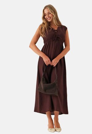Femme portant une robe longue marron sans manches tenant un sac à main marron foncé, avec de longs cheveux blonds et des chaussures plates couleur crème.