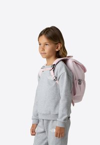 Light pink backpack with adjustable straps, rounded top, and front pocket, worn by a child in a gray sweatshirt and sweatpants.