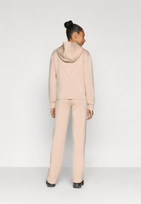 Μπεζ φούτερ με κουκούλα και αντίστοιχο παντελόνι φόρμας από μαλακό ύφασμα. Το φούτερ έχει ένα τριγωνικό λογότυπο στην πλάτη. Το μοντέλο στέκεται με casual στάση.