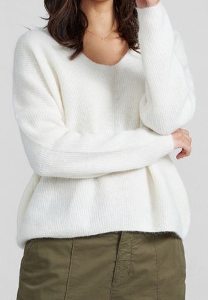 Woman wearing a white ribbed knit sweater with a V-neck and olive green pants, arms crossed in front against a plain background.