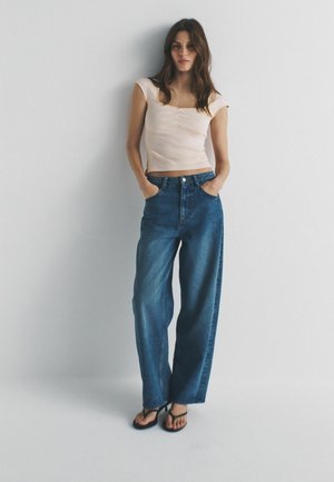 Hellrosa eng anliegendes Oberteil mit kurzen Kappenärmeln, kombiniert mit hoch taillierten, lockeren blauen Jeans aus Denim. Das Model steht mit den Händen in den Taschen.