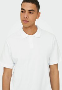 Hvid kortærmet polo-shirt lavet af tekstureret stof med klassisk krave og tre-knappet stolpe, med rullekanter på ærmerne.