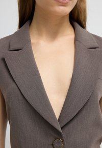 Hnědý oblekový blejzr s hlubokým véčkovým výstřihem, výraznými klopami a akcentem zlatého knoflíku. Hladká textura a strukturovaný design.