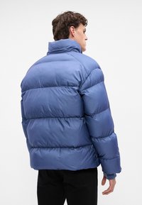 Giacca a vento blu con colletto alto, design trapuntato e polsini a coste. Presenta una vestibilità comoda e una texture esterna liscia e lucida.