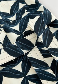 Tissu ceinturé avec un grand motif de feuilles bleu marine sur fond blanc, montrant des détails de couture et des passants de ceinture qui se chevauchent.