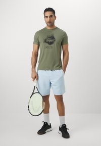 Olivno zelena majica s crnim grafičkim logotipom, svijetloplave kratke hlače, crne tenisice i drži tenisku reketa.
