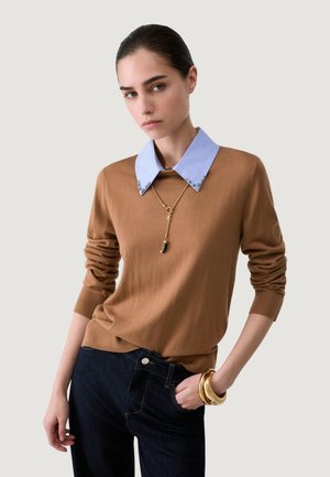 Joven mujer con un suéter marrón con un cuello azul adornado, vaqueros oscuros, collar de oro con colgante y varias pulseras de oro.
