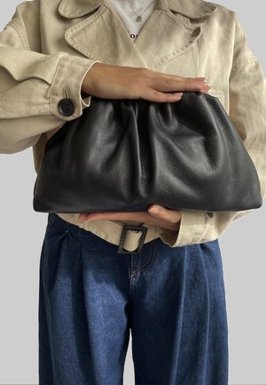 Borsa a mano in pelle nera con un design arricciato. Texture morbida e forma flessibile. Tenuta da una persona che indossa una giacca beige e pantaloni di denim.
