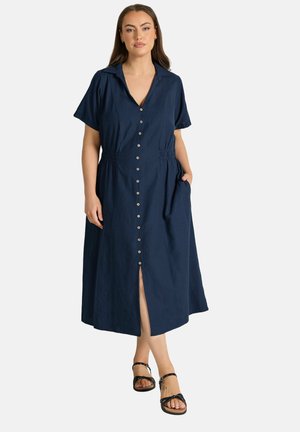 Vrouw draagt een midijurk in marineblauw met korte mouwen, een kraag, een voorste split en zwarte sandalen met bandjes, staand met één hand in haar zak.