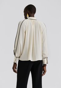 Μπλούζα με μακριά μανίκια σε ελεφαντίνι ύφασμα με μαύρο περίγραμμα κατά μήκος των μανικιών και γιακά. Χαλαρή εφαρμογή με λεπτομέρεια σούρας στην πλάτη.