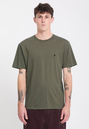 Ung mand med kort krøllet hår iført olivengrøn t-shirt og mørke bukser, står med neutralt ansigtsudtryk og synlige tatoveringer på begge arme.