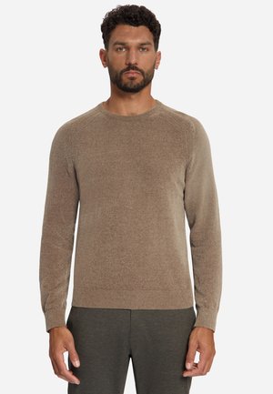 Man met kort krullend haar en baard, gekleed in een beige trui met lange mouwen en donkere broek tegen een effen lichte achtergrond.
