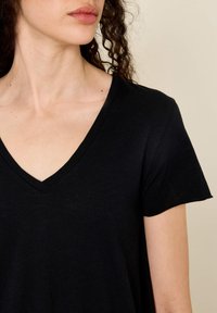 Woman with curly hair wearing a black V-neck short-sleeve shirt, shown from the neck to the upper arm against a beige background.