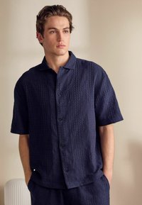 Camisa de botones de manga corta en azul marino con un patrón texturizado, tela tejida y cuello. Combinada con pantalones a juego, que muestran un ajuste relajado.