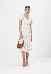 Robe florale blanche avec des manches courtes à volants, taille smockée et ourlet à volants. Associée à un sac bandoulière marron et des sandales à lanières.