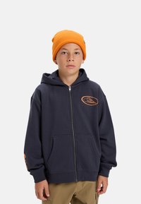 Navy zip-up hoodie with a front pocket and orange logo, paired with a bright orange knit beanie. The material appears soft and casual.