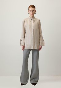 Bež majica na pruge oversized kroja s oštrim kragnom i širokim manžetama, u kombinaciji sa sivim trapezastim hlačama i smešnim cipelama s oštrim vrhom.