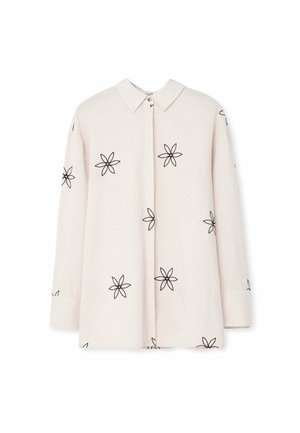 Camisa de manga larga color beige con cuello abotonado, presenta bordados de flores negras en el frente y las mangas. Textura de tela suave.