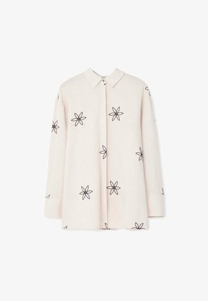Camisa de manga larga color beige con cuello abotonado, presenta bordados de flores negras en el frente y las mangas. Textura de tela suave.