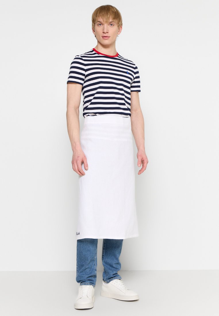 Un grembiule bianco indossato sopra jeans blu e sneakers bianche. Il modello indossa una maglietta a righe blu navy e bianche con un colletto rosso.