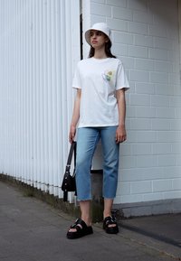 T-shirt branca com gráfico colorido, jeans cropped azul claro, sandálias plataformas pretas, e um chapéu bucket branco. Bolsa tiracolo preta.
