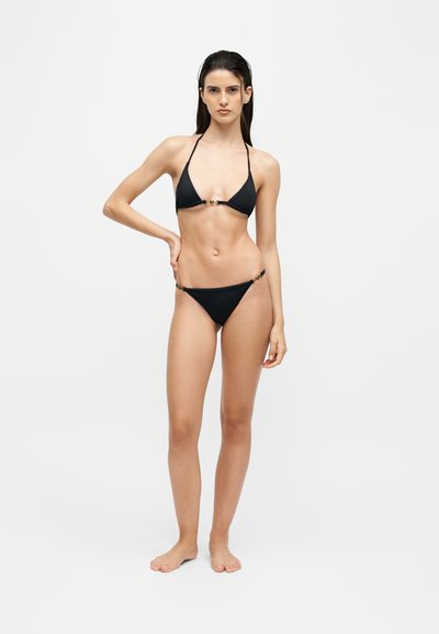 Jeune femme aux longs cheveux foncés, debout pieds nus, portant un bikini noir deux pièces sur un fond clair uni.