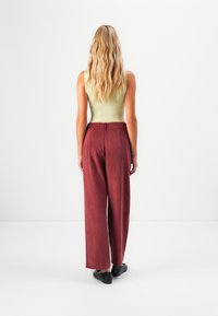 Persona con lunghi capelli biondi ondulati di spalle, indossante una top senza maniche beige, pantaloni larghi bordeaux e scarpe nere su uno sfondo bianco.