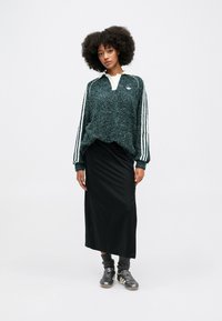Pulôver verde texturizado com colarinho branco, saia maxi preta e meias cinzentas combinadas com sapatos pretos e prateados; três riscas brancas nas mangas.