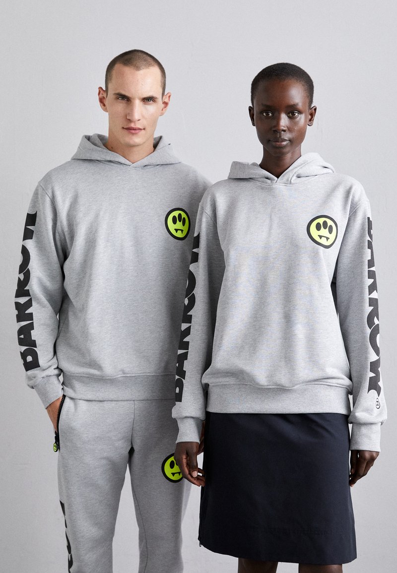Grå hettegensere med luer, med et grønt smilefjes-logo og svart tekst på ermer. I tillegg grå joggebukser og et svart skjørt.
