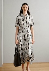 Printed button-up midi dress with short sleeves, featuring black, white, and gray geometric patterns. Model carries a black tote bag.
