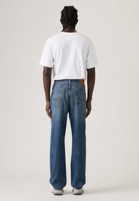 Μπλε τζιν παντελόνι με άνετη εφαρμογή, με τσέπες στο πίσω μέρος, δερμάτινη ετικέτα στη μέση και λευκό μπλουζάκι, απεικονιζόμενο από πίσω.