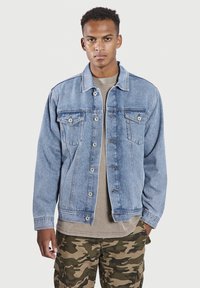 Lichtblauwe denim jack met twee borstzakken, zichtbare steeknaden, knoopsluiting en een ontspannen pasvorm. Draag het over een beige shirt en camouflagebroek.