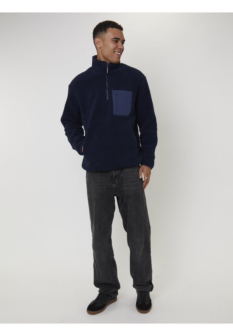 Jonge man die glimlacht, in een marineblauwe fleecepullover met borstzak, donkere spijkerbroek en zwarte sneakers, staande tegen een plain witte achtergrond.