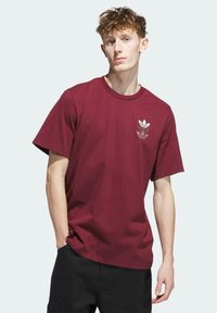 Camiseta de algodón burdeos de manga corta, con cuello redondo acanalado y un pequeño gráfico de logo blanco en el lado izquierdo del pecho.