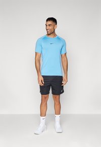 Blaues sportliches T-Shirt mit kurzen Ärmeln und einem schwarzen Nike-Logo, in Kombination mit schwarzen Shorts und weißen Turnschuhen, vor einem neutralen Hintergrund stehend.
