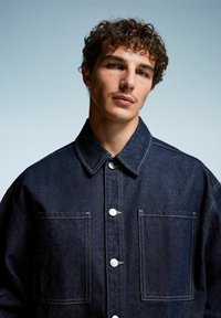 Giacca in denim scuro con chiusura a bottoni, colletto a punta e due tasche sul petto. Presenta cuciture a contrasto e una vestibilità rilassata.