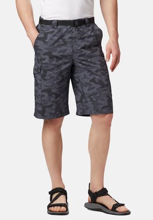 Uomo che indossa pantaloncini cargo mimetici grigio scuro con cintura, camicia bianca e sandali neri con cinturini, in piedi con le mani nelle tasche.