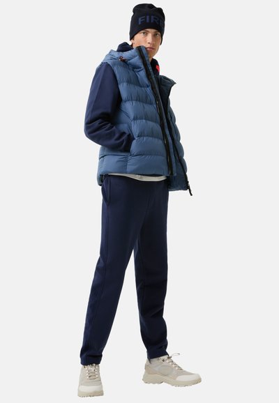 Blauwe puffer vest met horizontale stiksels, gedragen over een marineblauw sweatshirt en bijpassende broek. Lichte sneakers maken de look af.