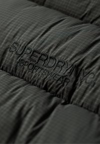 Chaleco de plumas negro con un patrón de cuadrícula. Presenta un logo bordado de "SUPERDRY NYC". El material parece ligero con una textura suave.