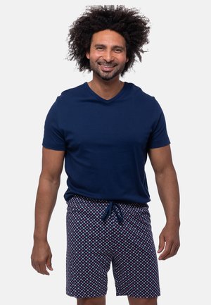 Uśmiechnięty mężczyzna o kręconych włosach, ubrany w granatową koszulę z krótkim rękawem oraz wzorzyste krótkie spodenki ze sznurkiem, stojący na jednolitym tle.