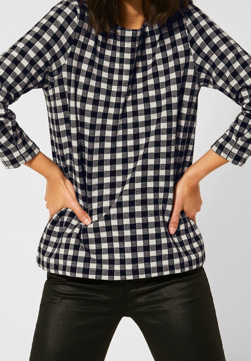 Navy and white gingham pattern top with three-quarter sleeves, featuring a round neckline and side pockets, worn over black pants.