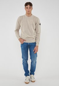 Giovane uomo in piedi, con un maglione beige, jeans blu e sneakers bianche con suole di gomma, davanti a uno sfondo bianco semplice.