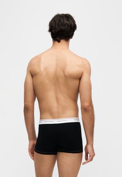 Homme torse nu de dos portant un boxer noir avec une ceinture blanche de la marque "DSQUARED2" sur un fond clair uni.