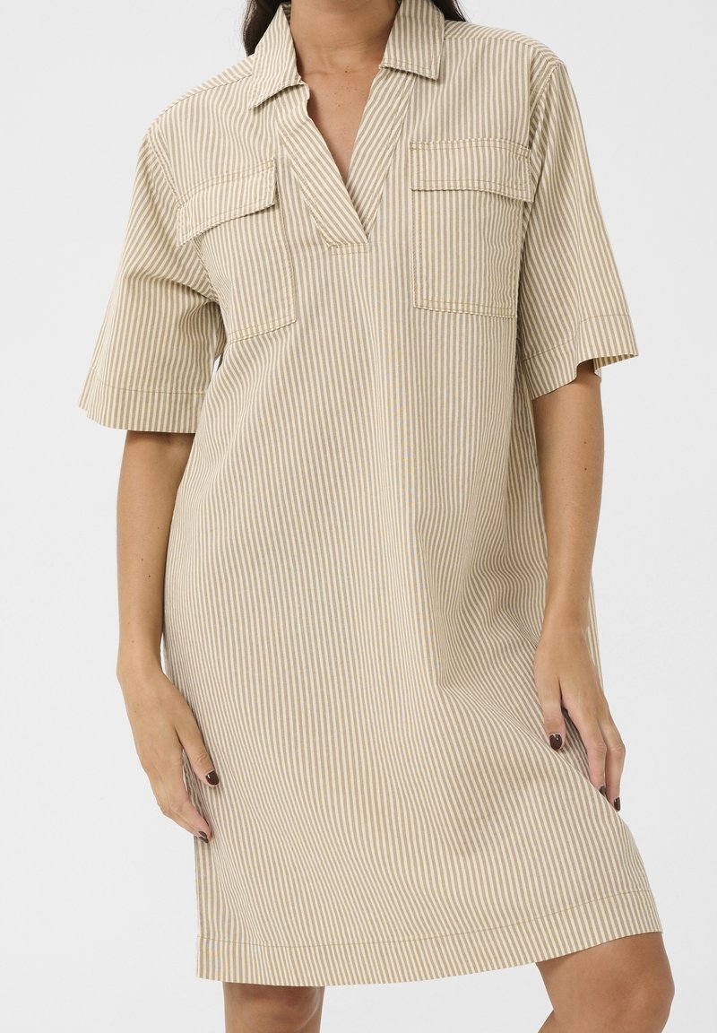 Femme portant une robe ample beige et blanche à rayures verticales, manches courtes, col en V et deux poches poitrine à rabats.