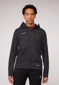 Zwarte softshell jas met een capuchon, voorzien van ritszakken, een gladde textuur en een aansluitend ontwerp. Merkopdruk op de borst en de onderrand.