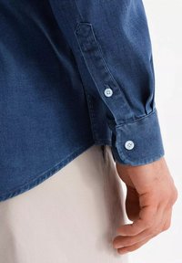 Camicia in denim blu scuro con polsini abbottonati, vestibilità sagomata, superficie liscia e orlo curvo. Dettagli di cucitura discreti sulle maniche.