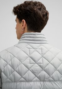 Giacca trapuntata grigia chiara con colletto alto e dettagli a motivo diamond sul retro. Tessuto liscio con righe di cuciture orizzontali.